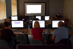 Uczestnicy Klubu Seniora siedzą przy stanowiskach w pracowni komputerowej podczas zajęć komputerowych, w tle ekran projekcyjny oraz trener zajęć prowadzący zajęcia