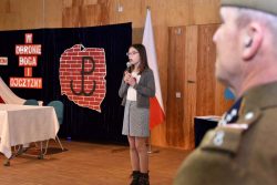 Na scenie sali stoi uczennica szkoły prezentujący piosenkę patriotyczną