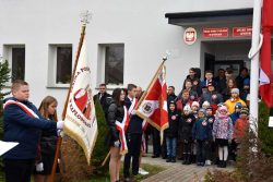 Przed wejściem do urzędu stoją uczniowie ze sztandarami szkół z terenu gminy Wyryki, w tle uczestnicy wydarzenia, przed nimi masz z opuszczoną flagą