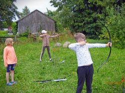 Uczestnicy spotkania stoją na trawniku, chłopiec i dziewczynka trzymają w dłoniach łuk celując do tarczy