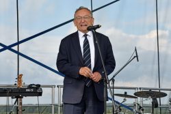Na zdjęciu Senator RP Józef Zając ubrany w granatową marynarkę, białą koszulę i krawat w granatowo-białe paski. Senator stoi na scenie, uśmiecha się i przemawia do mikrofonu.