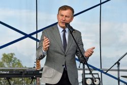 Na zdjęciu Poseł na Sejm Krzysztof Grabczuk ubrany w szarą marynarkę w drobną kratkę, jasnoniebieską koszulę i ciemny krawat. Poseł na Sejm stoi na scenie, przemawia do mikrofonu, gestykulując obiema rękami.