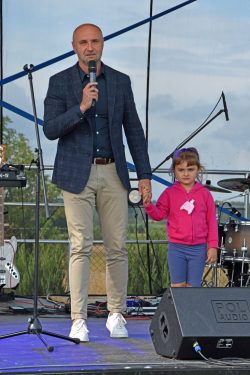 Na zdjęciu widać mężczyznę i małą dziewczynkę stojących na scenie. Mężczyzna, ubrany w granatową marynarkę, ciemną koszulę i beżowe spodnie, trzyma w prawej ręce mikrofon i patrzy w stronę obiektywu. Dziewczynka, stojąca po jego prawej stronie i trzymająca go za rękę,