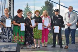Grupa siedmiu osób stojących na scenie. Wszystkie osoby trzymają wręczone podziękowania od Wójta Gminy.