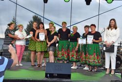 Grupa kobiet stojących na scenie. Niektóre z nich trzymają w rękach dyplomy i nagrody pieniężne. W tle widoczne są elementy sceny.