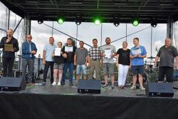 Grupa dziesięciu osób stojących na scenie. W centralnej części sceny, pod zielonymi reflektorami, znajduje się osoby, które trzymają w rękach dyplomy i nagrody pieniężne. Po lewej stronie stoi mężczyzna z mikrofonem, a obok niego drugi mężczyzna w okularach przeciwsłonecznych.