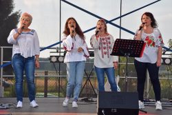 Zespół Mołodyczki, cztery kobiety stojące na scenie, śpiewające do mikrofonów. Wszystkie są ubrane w białe bluzki z ludowymi haftami oraz dżinsy.
