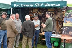 Grupa mężczyzn stoi pod zielonym namiotem z napisem „Koło Łowieckie Żbik Warszawa”. W tle namiotu widoczne są wypchane zwierzęta i skóry.