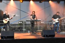 Zespół muzyczny nBlue składający się z trzech osób występujących na scenie. Wszyscy ubrani są na ciemno. Po lewej stronie stoi mężczyzna z okularami, grający na białej gitarze elektrycznej. Na środku sceny stoi kobieta grająca na keyboardzie i śpiewająca do mikrofonu. Po prawej stronie stoi mężczyzna grający na białej gitarze basowej.