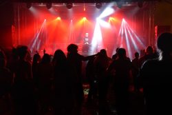 Na zdjęciu widać scenę, na której DJ Ario gra na sprzęcie, oświetlony jasnymi, białymi i czerwonymi reflektorami, które tworzą dynamiczne snopy światła i dymu. Przed sceną znajduje się tłum ludzi, których sylwetki są widoczne w ciemności. Ludzie są zwróceni w stronę sceny, niektórzy mają uniesione ręce.