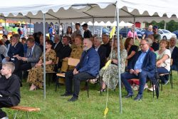 Grupa osób siedzi na krzesłach pod białym namiotem. W tle widać więcej ludzi i samochody.