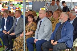 Grupa osób siedzi na krzesłach pod białym namiotem. W tle widać innych ludzi i zarysy budynków.