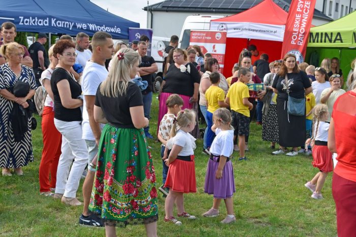 Na zdjęciu widać grupę osób, w tym dorosłych i dzieci, zgromadzonych na trawiastym terenie. W tle widoczne są namioty – niebieski, na którym częściowo widnieje napis „Zakład Karny we Włodawie”, oraz czerwony z napisem „Poleskie Psy Ratownicze”.