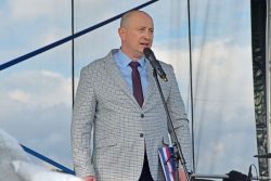 Na zdjęciu Wójt Gminy Wyryki Bernard Błaszczuk, ubrany w elegancką, jasną marynarkę w kratę, niebieską koszulę i bordowy krawat. Stoi na scenie i mówi do mikrofonu.