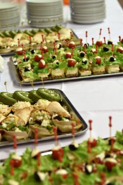 Zbliżenie na stół z cateringiem, na którym znajdują się tace z różnorodnymi przekąskami typu finger food i koreczkami. W górnej części widoczne są stosy białych talerzy.