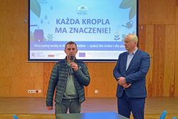 Wójt Gminy Wyryki oraz wykonawca stoją na scenie przed ekranem projekcyjnym z napisem w języku polskim: "KAŻDA KROPLA MA ZNACZENIE!".