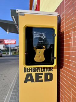 Paczkomat InPost z zamontowanym defibrylatorem AED w żółtej obudowie, stojąca przy sklepie spożywczym Delikatesy w słoneczny dzień.