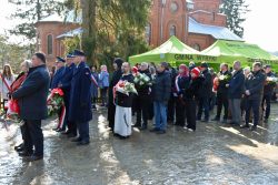 Uczestnicy uroczystości patriotycznej w Suchawie stoją przed kościołem, trzymając wieńce i wiązanki kwiatów; w tle widoczny namiot Gminy Wyryki.