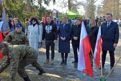 Uczestnicy uroczystości patriotycznej stoją przy pomniku, a żołnierz składa kwiaty; w tle widoczne polskie flagi.