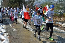 Uczestnicy biegu „Tropem Wilczym” – dzieci i dorośli – biegną wspólnie leśną drogą, ubrani w okolicznościowe koszulki.