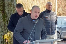 Wójt Gminy Wyryki w czarnym płaszczu przemawia do mikrofonu na przezroczystej mównicy.