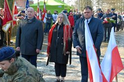Uczestnicy uroczystości patriotycznej stoją przy pomniku, a żołnierz składa kwiaty; w tle widoczne polskie flagi.