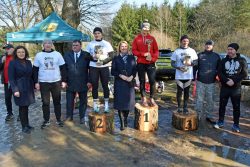 Treść wiadomości Gemini Uczestnicy biegu "Tropem Wilczym" na podium zrobionym z pni drzew. Zwyciężczyni w czerwonej bluzie stoi na najwyższym stopniu, trzymając puchar. Obok niej znajdują się zdobywczynie drugiego i trzeciego miejsca oraz organizatorzy i goście honorowi. W tle widać namiot z logiem wydarzenia i las.