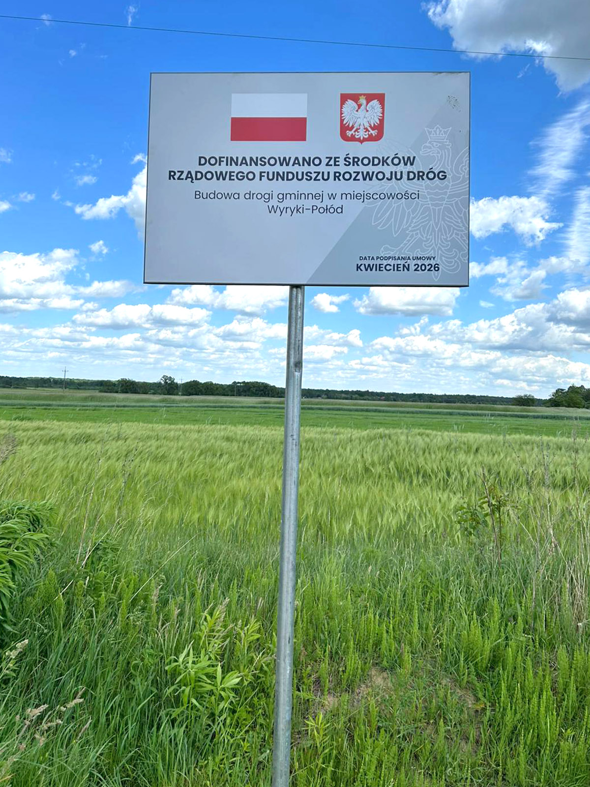 Tablica informacyjna o dofinansowaniu budowy drogi gminnej w miejscowości Wyryki-Połód ustawiona przy polu, na tle błękitnego nieba z chmurami.